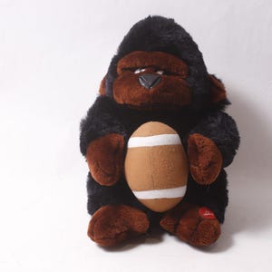 May include: A soft toy gorilla with black fur and brown facial features, hands, and feet. It is holding a brown American football with white stripes. A red tag is attached to its foot.