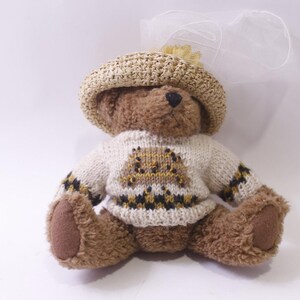 Pode incluir: Um urso de pelúcia marrom vestindo um suéter de malha bege com um design geométrico e um chapéu combinando. O urso tem um nariz preto e está sentado. Um véu branco está preso ao chapéu.