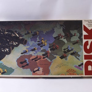 May include: A vintage Risk board game box with a world map and a cannon on the front. The box is red and white with the word "RISK" in large, bold letters.