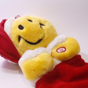 May include: A plush yellow smiley face with a winking eye, black smile, and black eyes. It is wearing a red Santa hat and coat with white trim. A red button with the words "PRESS HERE" is on the arm.