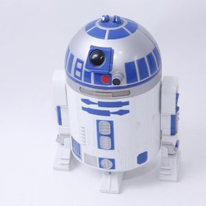 May include: A white and blue R2-D2 toy robot with a silver dome head. The robot has blue and silver details, including panels and lights. The toy is a Star Wars collectible.