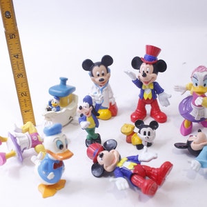 May include: A collection of Disney character figurines, including Mickey Mouse, Donald Duck, and Goofy. The figures are brightly colored and in various poses, with a yellow ruler in the background.