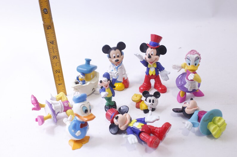 May include: A collection of Disney character figurines, including Mickey Mouse, Donald Duck, and Goofy. The figures are brightly colored and in various poses, with a yellow ruler in the background.