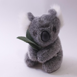 May include: A plush gray koala bear with white tufts of fur on its ears, holding a green leaf. The koala has a black nose and is sitting upright. The toy is designed to resemble a koala.