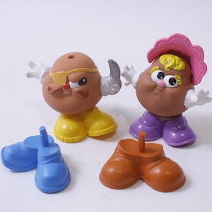 Könnte beinhalten: Zwei Mr. Potato Head Spielzeuge, eines mit braunem Körper, gelben Schuhen und einer schwarzen Augenklappe, das andere mit braunem Körper, lila Schuhen und einem rosa Hut. Beide Spielzeuge haben austauschbare Teile.