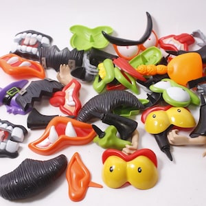 Puede incluir: Una colección de piezas de juguetes de plástico con temática de Halloween, que incluyen dientes, ojos, orejas y sombreros de bruja. Las piezas son de varios colores como naranja, verde, negro y morado, lo que sugiere un tema festivo.