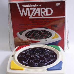 May include: A vintage Waddingtons Wizard electronic game. The game features a white, blue, yellow, and red base with a black playing surface. The box is red with the game's name and features. The game plays 4 electronic games.
