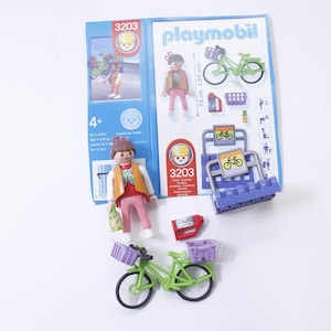 May include: Playmobil 3203 set featuring a girl figure, a green bicycle with a basket, and a purple bike rack with a sign that reads "Playmobil 3203".