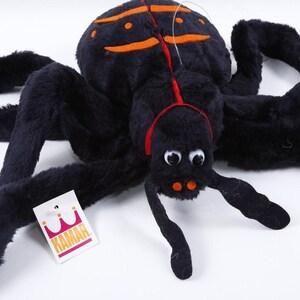 May include: A plush black spider toy with eight long legs, googly eyes, and orange markings on its back. The spider features a red line down its center and two orange spots near its mouth. A white tag with a pink crown logo and the text KAMAR is attached.