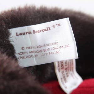 May include: A white tag with brown text that reads "Lauren Bearcall TM 1983 All Rights Reserved North American Bear Company, Inc. Chicago, NY (312) 943-4061".