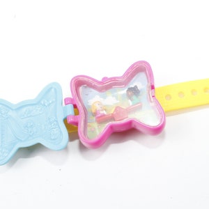 May include: A pink and blue butterfly-shaped plastic wristwatch with a clear window showing two small dolls playing on a swing set. The watch has a yellow adjustable band with five holes.