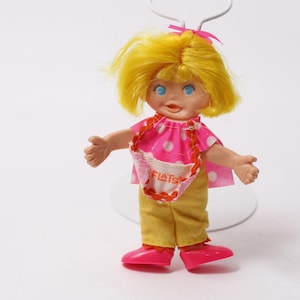 Puede incluir: Muñeca vintage con cabello amarillo, ojos azules y un lazo rosa. La muñeca viste una camisa rosa con lunares, pantalones amarillos y zapatos rosas. Un pequeño delantal con la palabra "FIATS" es visible.