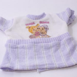 Puede incluir: Un atuendo de muñeca vintage. La parte superior blanca tiene un gráfico "HUGGA BUNCH" y personajes de dibujos animados. El atuendo incluye una falda lavanda con rayas verticales y mangas a juego.
