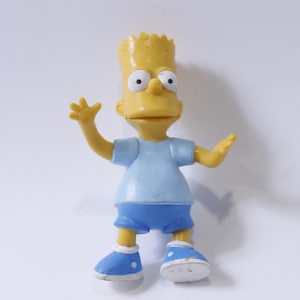 May include: A yellow plastic figurine of Bart Simpson from The Simpsons. He is wearing a blue shirt and blue shorts and has a yellow head with black hair. He is waving with his right hand and has a surprised expression on his face.