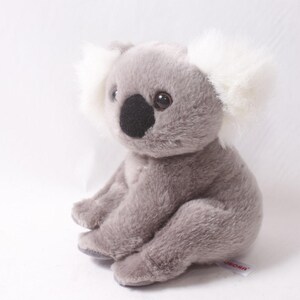 May include: A soft toy koala bear, grey in colour, with fluffy white ears and a black nose. The koala is in a seated position against a white backdrop. The toy has large, round eyes.