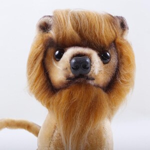 May include: A plush lion toy with a tan body and a brown mane. The lion has large, black eyes and a black nose. The tail is visible in the lower left corner. The toy is set against a white background.