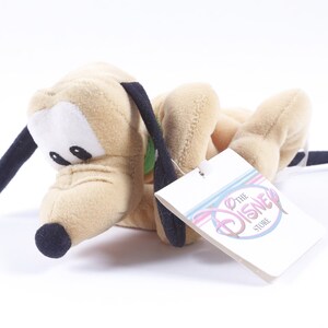 Puede incluir: Un perro de peluche marrón claro con orejas negras y nariz negra. El perro está acostado de lado con la cabeza apoyada en las patas. El perro lleva un collar verde y una etiqueta que dice "The Disney Store".