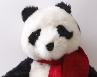 May include: A white and black plush panda bear wearing a red knitted scarf. The panda has black eyes and a black nose.