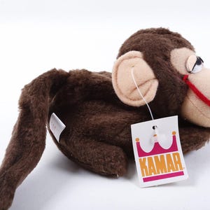 May include: A brown plush monkey toy with a beige face, ears, and a small pink nose. It features a red stitched mouth and a white tag attached to its ear with the text KAMAR printed in orange letters above a pink crown graphic on a white background.