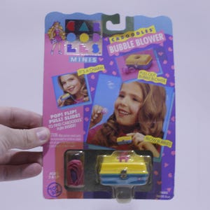 May include: A vintage Caboodles Bubble Blower toy package. The packaging is pink and purple, featuring images of a girl and the toy. The bubble blower is yellow and blue. The text on the package reads "Caboodles Bubble Blower".