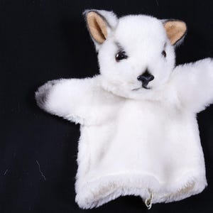 May include: A white plush hand puppet shaped like a small animal with pointed ears, dark brown inner ear accents, black eyes, and a black nose. The puppet features soft, fuzzy white fur covering the head and arms, with a simple open base design.