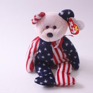 May include: A patriotic plush bear with a cream-colored head and a body featuring red and white stripes and blue with white stars. The bear has a red and white striped bow and a tag.