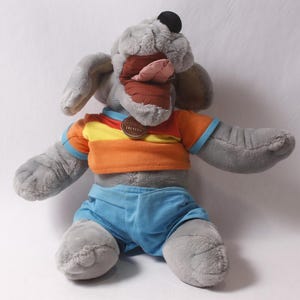 Puede incluir: Un juguete de peluche gris con forma de perro con la boca abierta, que lleva una camiseta a rayas naranja y amarilla, pantalones cortos azules y una pequeña etiqueta circular. El perro tiene orejas caídas y una nariz negra.