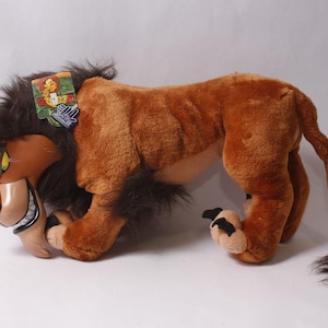 May include: A plush toy of a lion with brown fur, a dark brown mane, and a molded plastic face featuring green eyes and a wide grin. The character has a long tail with a dark tuft at the end and a small rectangular tag attached to the mane.