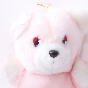 Peut inclure: Un ours en peluche rose avec un visage blanc et des yeux rouges. L'ours porte un halo doré.
