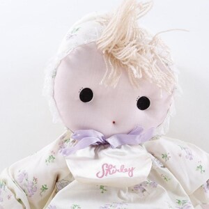 May include: A soft doll with a light pink face, large black eyes, and a small red nose. The doll has light blonde yarn hair and wears a floral print dress with a matching bonnet. A bib with the name "Shirley" is attached.