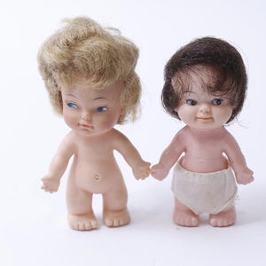 May include: Two plastic baby dolls, one with blonde hair and blue eyes and the other with brown hair and brown eyes. Both dolls are wearing white diapers.