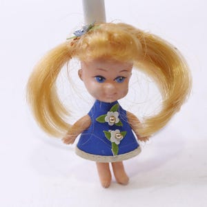 May include: A vintage doll with blonde pigtails and blue eyes. The doll wears a blue dress with white flower and green leaf accents. The doll's dress has a beige trim. The doll is standing on a white surface.