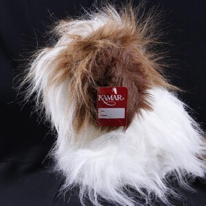 May include: A plush toy dog with long, shaggy white and brown fur. The toy features a rectangular red tag attached to the front with the white text KAMAR printed on it, along with blank spaces for size and stock information below the brand name.