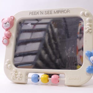 May include: A cream-colored baby mirror with the text "PEEK 'N SEE MIRROR". The mirror has a rectangular shape with rounded corners and is decorated with pink and blue teddy bears. It also features three colorful beads.