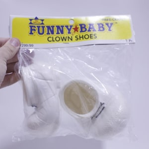 May include: A pair of white clown shoes in a clear plastic bag. The shoes have a rounded shape and a small black strap detail. The packaging has a yellow label with the text "FUNNY BABY CLOWN SHOES".