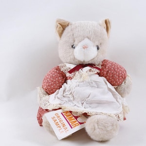 May include: A plush cat toy with beige fur, a white muzzle, and a pink nose. The cat wears a red dress with a small white floral pattern, a white apron with lace trim, and a dark red ribbon at the neck. A tag reads HAMAR ORIGINAL SAMPLE.