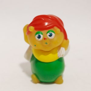 Könnte beinhalten: Eine gelbe und grüne Plastikfigur einer Raupe mit rotem Kopf und großen Augen. Die Raupe lächelt und hat einen weißen Flügel auf dem Rücken.