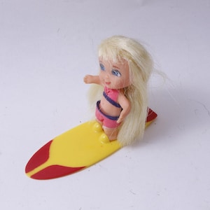 Peut inclure: Une petite poupée aux longs cheveux blonds, vêtue d'un maillot de bain rose avec des accents bleus, se tient sur une planche de surf jaune et rouge. La poupée a les yeux bleus et est posée comme si elle surfait. La planche de surf mesure environ 13 cm de long.