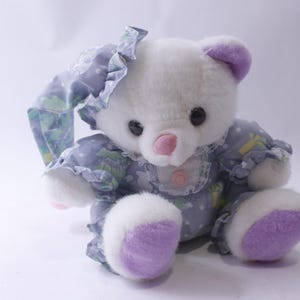 Op de afbeelding: Een witte pluche teddybeer die een lavendel en wit gepatroneerd slaappak draagt met een paarse strik. De beer heeft een roze neus en paarse pootkussens.