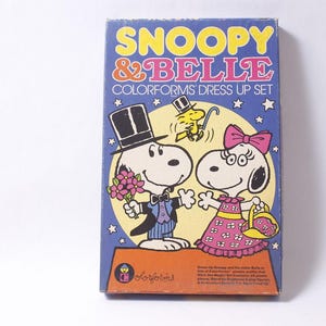 May include: Vintage Colorforms dress-up set featuring Snoopy and Belle. The box has a blue background with Snoopy in a tuxedo and Belle in a pink dress. The text "Snoopy & Belle" is in yellow and pink.