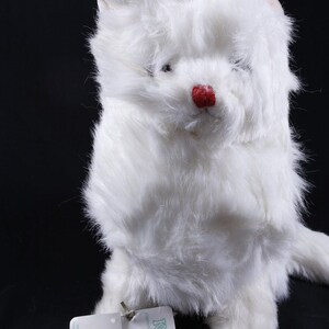 May include: A plush white cat toy with long, soft synthetic fur, pointed ears, and a small, round, bright red nose. A rectangular white paper tag is attached to the front, featuring the text Kicks, Angora, and the number 5588 in blue ink.