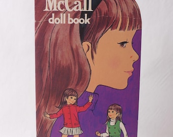 Puede incluir: Un libro de muñecas "Betsy McCall" vintage con una gran ilustración del perfil de una niña en tonos marrones, morados y rosas. Se presentan dos ilustraciones de muñecas, una con abrigo y falda rojos, la otra con un conjunto verde. El título del libro está en blanco.