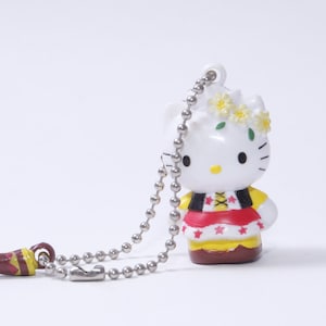 Peut inclure: Porte-clés Hello Kitty blanc avec une robe rouge et jaune, une couronne de fleurs et un charme de corne brune.