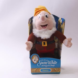 May include: A Sneezy soft toy from Disney's Snow White and the Seven Dwarfs, presented in its original packaging. The dwarf features a white beard, a yellow hat, and a brown outfit. The packaging is blue with the movie title.