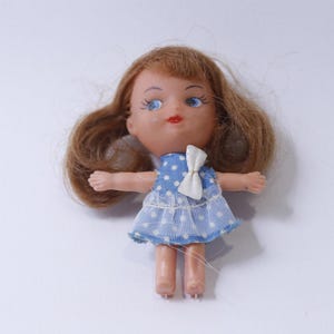May include: A small plastic doll with brown hair, blue eyes, and a blue and white polka dot dress. The doll is wearing a white bow.