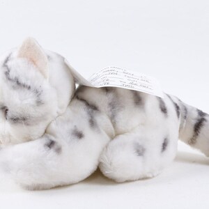 May include: A plush white cat with gray spots and stripes, lying on its side with its head tucked in. The stuffed animal has a tag attached to its back. The cat's fur is soft and fluffy, with a realistic design.