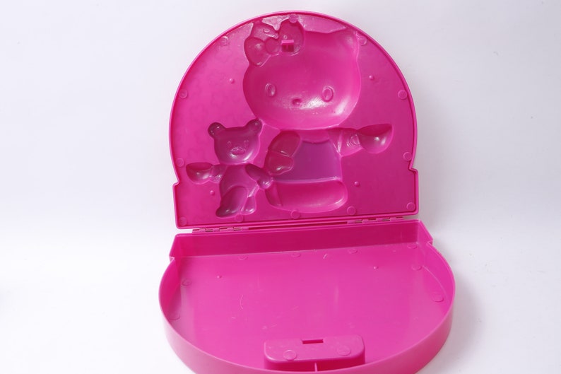 May include: Pink plastic Hello Kitty lunchbox with a molded image of Hello Kitty and a bear. The lunchbox is open and the inside is empty.