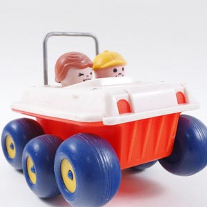 Puede incluir: Un coche de juguete vintage con carrocería roja, techo blanco y ruedas azules. Dos pequeñas figuras con pelo castaño y un sombrero amarillo están sentadas dentro. El juguete tiene una barra antivuelco plateada y detalles amarillos en las ruedas.