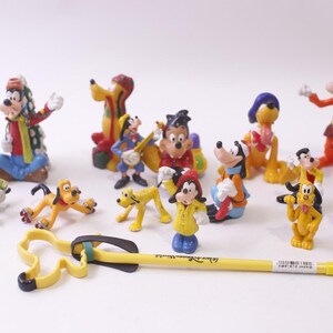 Pode incluir: Uma coleção de figuras de personagens da Disney, incluindo Mickey Mouse, Minnie Mouse, Pateta e Pluto. As figuras são coloridas e em várias poses. Uma caneta amarela com um design preto e azul está em primeiro plano.