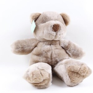 May include: A plush teddy bear with soft, light brown fur, rounded ears, and a darker brown stitched nose. The stuffed animal is in a seated position with outstretched arms and legs, featuring a small light green tag attached to its left ear.